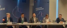 5 osób siedzących za stołem w sali konferencyjnej, w tle banery z logotypami i napisami Fundusze Europejskie