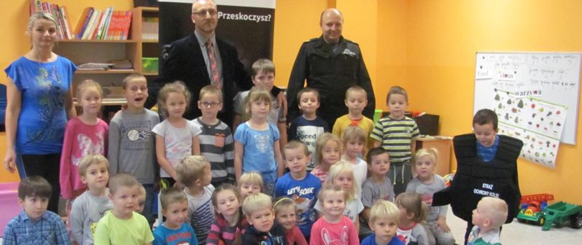 Grupa dzieci siedzi i stoi na dywanie, za nimi kobieta i dwóch mężczyzn. 