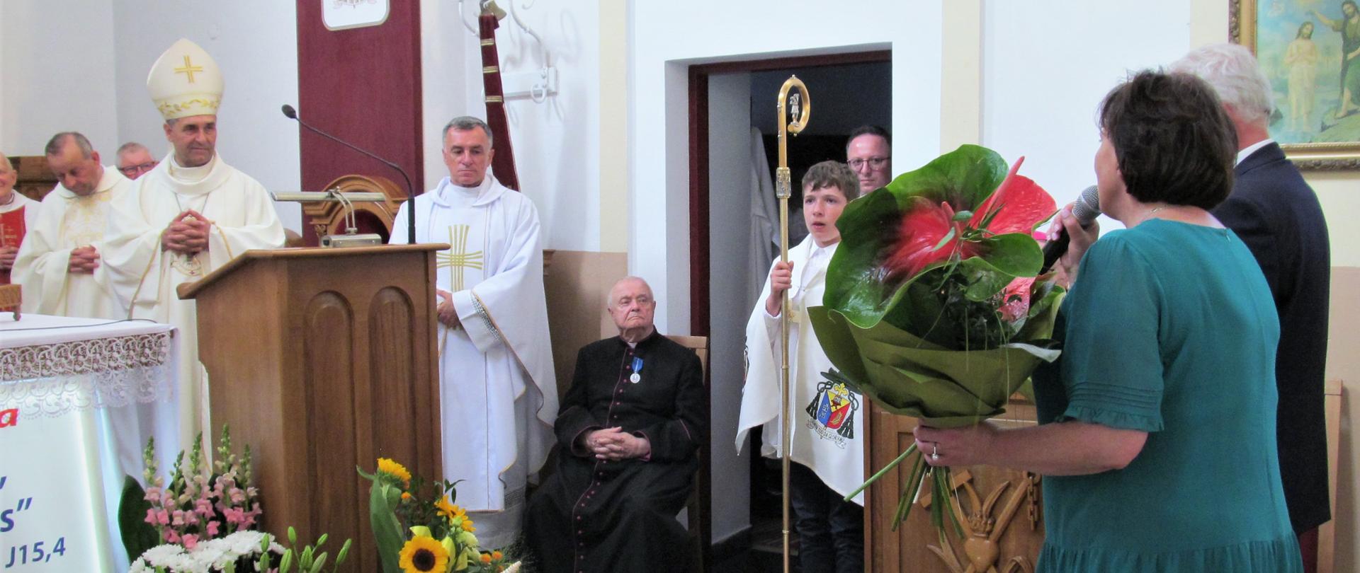 Na zdjęciu widoczny fragment prawej strony ołtarza. Po środku fotografii na drewnianym krześle siedzi ksiądz Jubilat, na którego piersi widnieje złoty medal. Po lewej stronie widać ks. bpa. dr Piotra Sawczuka, który kieruje wzrok na wchodzącą na ołtarz starostę Elżbietę Sadowską oraz senatora RP Waldemara Kraskę.