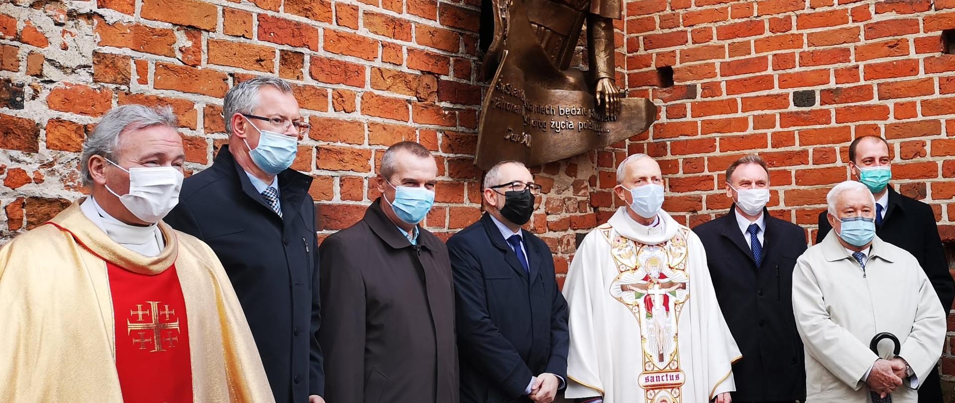 Zdjęcie zrobione podczas uroczystego odsłonięcia okna papieskiego w kościele farnym w Przasnyszu, Od strony lewej stoją: Proboszcz parafii Świętego Stanisława Kostki w Przasnyszu ksiądz prałat Andrzej Maciejewski, Radny Rady Powiatu Przasnyskiego Jarosław Szmulski, Wicestarosta Powiatu Przasnyskiego Waldemar Trochimiuk, Starosta Przasnyski Krzysztof Bieńkowski, Biskup Płocki Piotr Libera, Przewodniczący Rady Miejskiej w Przasnyszu Wojciech Długokęcki, rzeźbiarz Aleksander Porożniuk, Burmistrz Przasnysza Łukasz Chrostowski. Nad zebranymi na zdjęciu panami widnieje popiersie Świętego Jana Pawła II z płaskorzeźbą Świętego Stanisława Kostki, która to kompozycja została umieszczona w jednej z nisz przy głównym wejściu do kościoła.