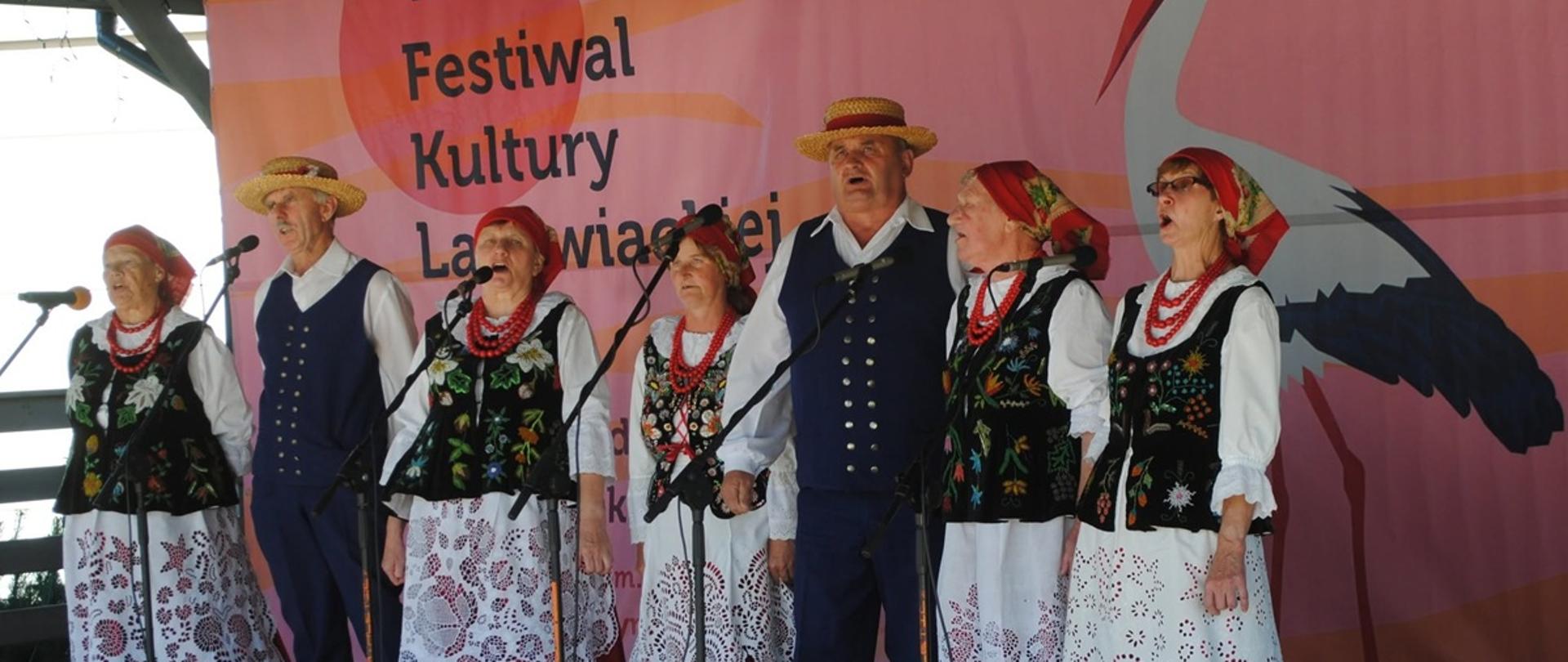Grupa osób w tradycyjnych strojach ludowych stoi na scenie i śpiewa do mikrofonów podczas festiwalu folklorystycznego. Kobiety mają białe haftowane spódnice, czarne haftowane kamizelki, czerwone chusty i korale, a mężczyźni ubrani są w ciemne stroje z kapeluszami oraz czerwonymi akcentami. W tle znajduje się duży baner z grafiką bociana i napisem dotyczącym festiwalu kultury ludowej.