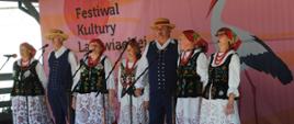 Grupa osób w tradycyjnych strojach ludowych stoi na scenie i śpiewa do mikrofonów podczas festiwalu folklorystycznego. Kobiety mają białe haftowane spódnice, czarne haftowane kamizelki, czerwone chusty i korale, a mężczyźni ubrani są w ciemne stroje z kapeluszami oraz czerwonymi akcentami. W tle znajduje się duży baner z grafiką bociana i napisem dotyczącym festiwalu kultury ludowej.