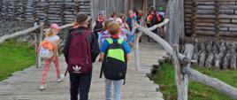 dzieci z plecakami na plecach idące po drewnianej kładce w kierunku bramy w drewnianym ogrodzeniu