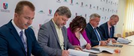 czterech mężczyzn i jedna kobieta siedzą przy stole i podpisują plik kartek 