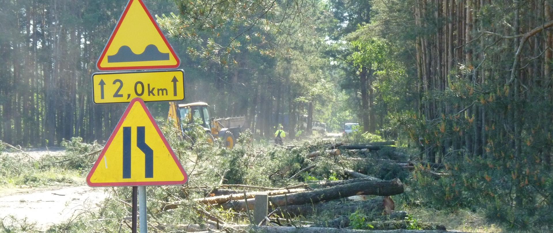Znak drogowy wyboje 2km, zwężenie prawostronne drogi. Za znakiem leżą powalone drzewa