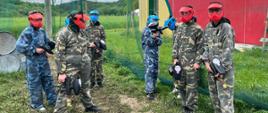 Grupka uczniów ubrana w specjalne stroje i maski na twarzach stoi na terenie przeznaczonym do gry w paintballa, trzymając w rękach specjalne pistolety do tej gry.