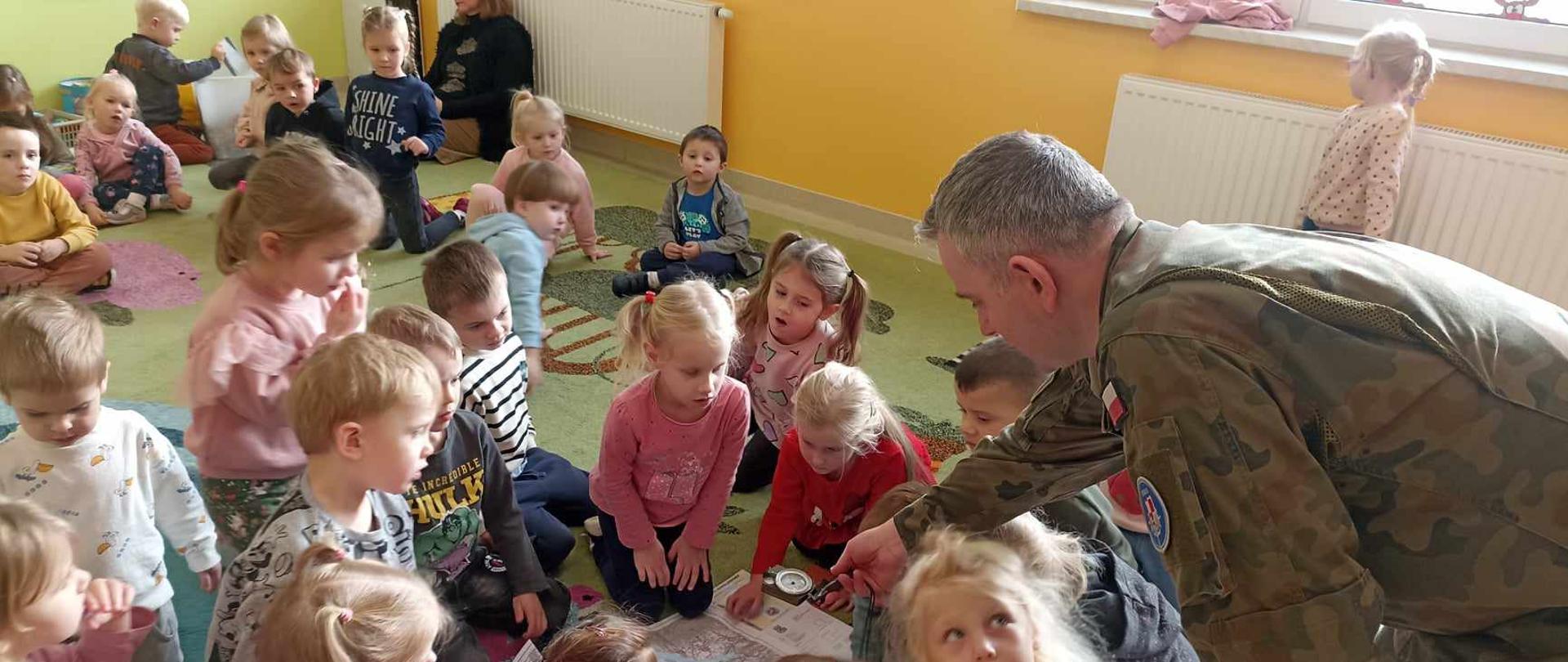 Żołnierz pokazuje siedzącym na dywanie dzieciom mapę i kompas