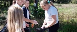 Cztery dziewczynki wrzucają znalezione wzdłuż drogi śmieci do dużego czarnego worka