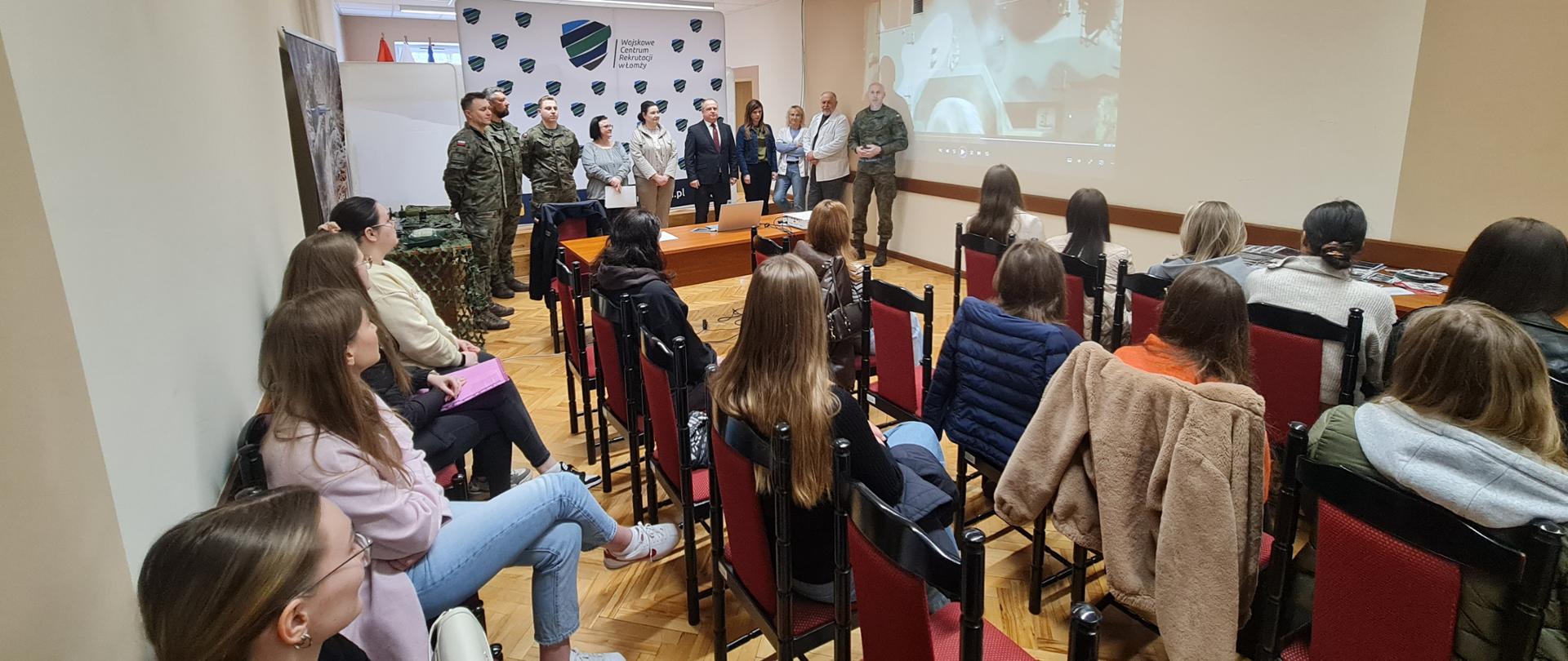 Zdjęcie przedstawia spotkanie informacyjne w jasnej, podłużnej sali konferencyjnej. Na pierwszym planie i w rzędach krzeseł siedzi grupa młodych osób, głównie tyłem lub bokiem do obiektywu, uważnie słuchając prelegentów.
W głębi pomieszczenia, pod jasną ścianą, stoi grupa około dziesięciu osób. Wśród nich znajdują się umundurowani żołnierze oraz osoby w strojach formalnych i cywilnych. Jeden z mężczyzn w mundurze stoi najbliżej środka i przemawia. Po lewej stronie, za grupą oficjeli, widoczna jest biała ścianka reklamowa z logo i napisem „Wojskowe Centrum Rekrutacji w Łomży”.
Po prawej stronie prelegentów, bezpośrednio na ścianie, wyświetlany jest z rzutnika materiał wideo przedstawiający ujęcia związane z wojskiem. Pomiędzy pierwszą linią krzeseł a występującymi osobami ustawiono drewniane biurko z otwartym laptopem. Sala ma jasne ściany, kasetonowy sufit z podłużnymi lampami oraz parkietową podłogę.