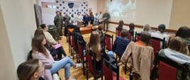 Zdjęcie przedstawia spotkanie informacyjne w jasnej, podłużnej sali konferencyjnej. Na pierwszym planie i w rzędach krzeseł siedzi grupa młodych osób, głównie tyłem lub bokiem do obiektywu, uważnie słuchając prelegentów.
W głębi pomieszczenia, pod jasną ścianą, stoi grupa około dziesięciu osób. Wśród nich znajdują się umundurowani żołnierze oraz osoby w strojach formalnych i cywilnych. Jeden z mężczyzn w mundurze stoi najbliżej środka i przemawia. Po lewej stronie, za grupą oficjeli, widoczna jest biała ścianka reklamowa z logo i napisem „Wojskowe Centrum Rekrutacji w Łomży”.
Po prawej stronie prelegentów, bezpośrednio na ścianie, wyświetlany jest z rzutnika materiał wideo przedstawiający ujęcia związane z wojskiem. Pomiędzy pierwszą linią krzeseł a występującymi osobami ustawiono drewniane biurko z otwartym laptopem. Sala ma jasne ściany, kasetonowy sufit z podłużnymi lampami oraz parkietową podłogę.