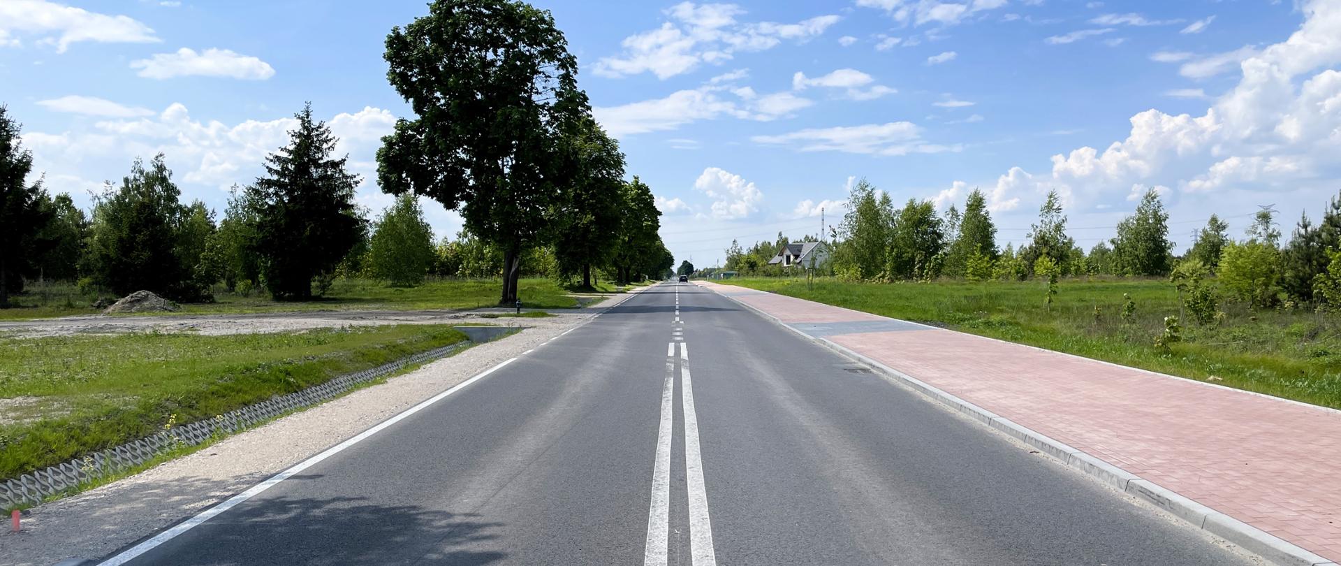 Droga asfaltowa, po prawej stronie czerwono-granatowy chodnik, po lewej stronie rów. W oddali widać drzewa i budynki.