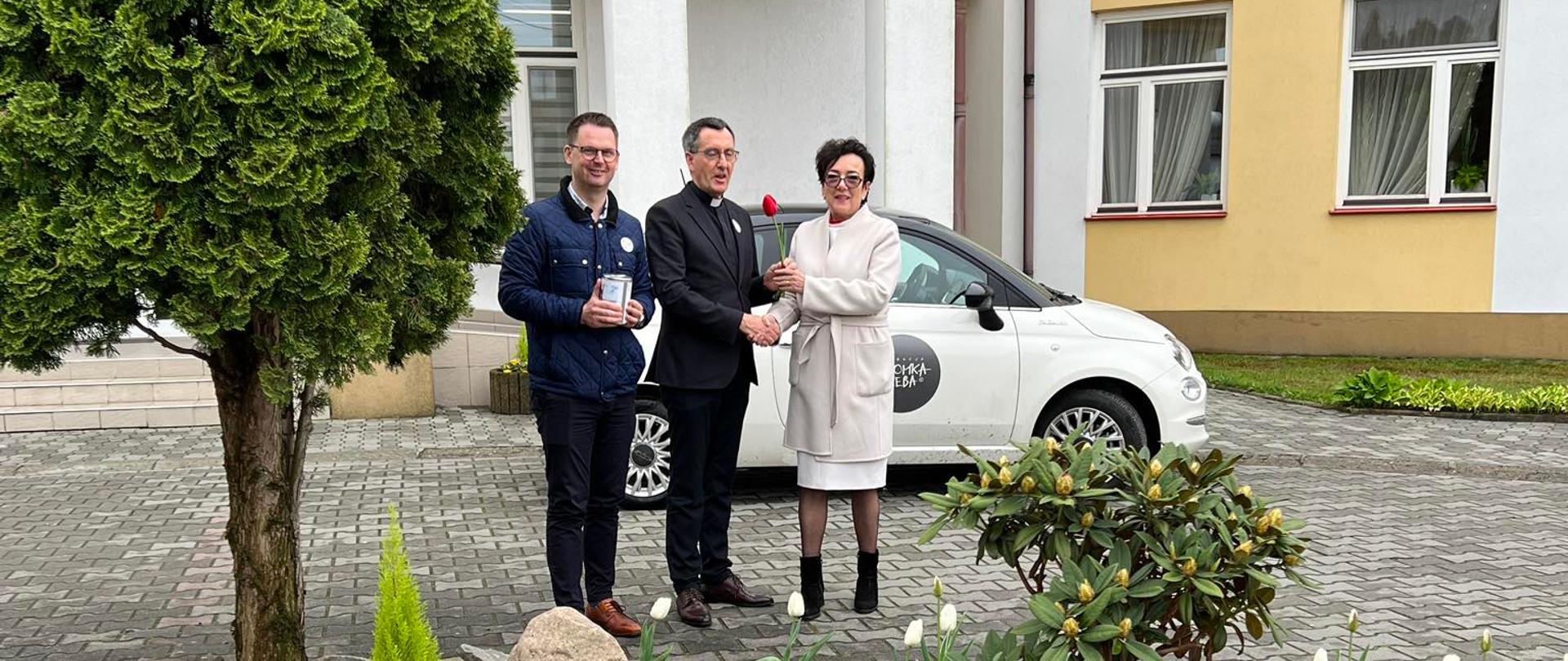 Zdjęcie przedstawia dwóch mężczyzn oraz kobietę. Od lewej: Tomasz Banek- wójt gminy Wietrzychowice, Ks. Marek Kogut-dyrektor Ośrodka "Caritas" w Jadownikach Mokrych oraz Anna Czech- prezes fundacji Kromka Chleba, stoją przed budynkiem Ośrodka "Caritas".