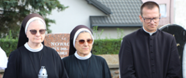 Dwie siostry zakonne w habitach oraz stojącego obok nich księdza ze splecionymi dłońmi. Jedna z zakonnic trzyma w rękach znicz ozdobiony biało-czerwoną wstęgą.