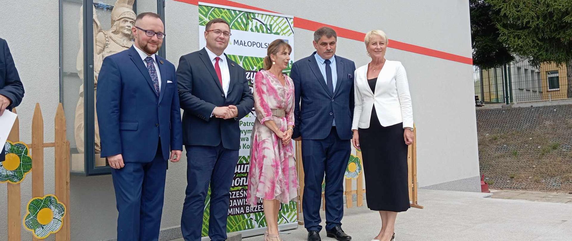 Pięcioro uczestników uroczystości pozuje stojąc pod szarą ścianą remizy strażackiej, za nimi drewniana figura św. Floriana i rollup promujący Małopolskę