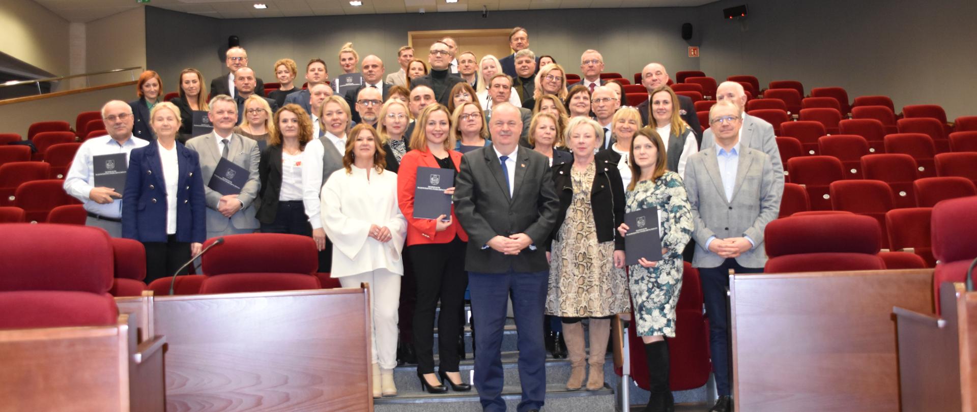 Na fotografii duża grupa osób. Osoby znajdują się na auli. Dookoła nich czerwone fotele. Na pierwszym planie widać brązowe biurka 