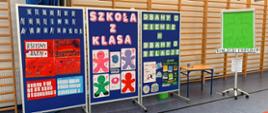 Po lewej stronie widać niebieską tablicę składającą się z trzech skrzydeł. Na niej wiszą plakaty wykonane przez dzieci oraz kolorowe napisy. W tle widać drabinki gimnastyczne. Po prawej stronie stoi stolik szkolny, Obok niego po prawej stronie znajduje się tablica, na którym wisi zielony brystol.