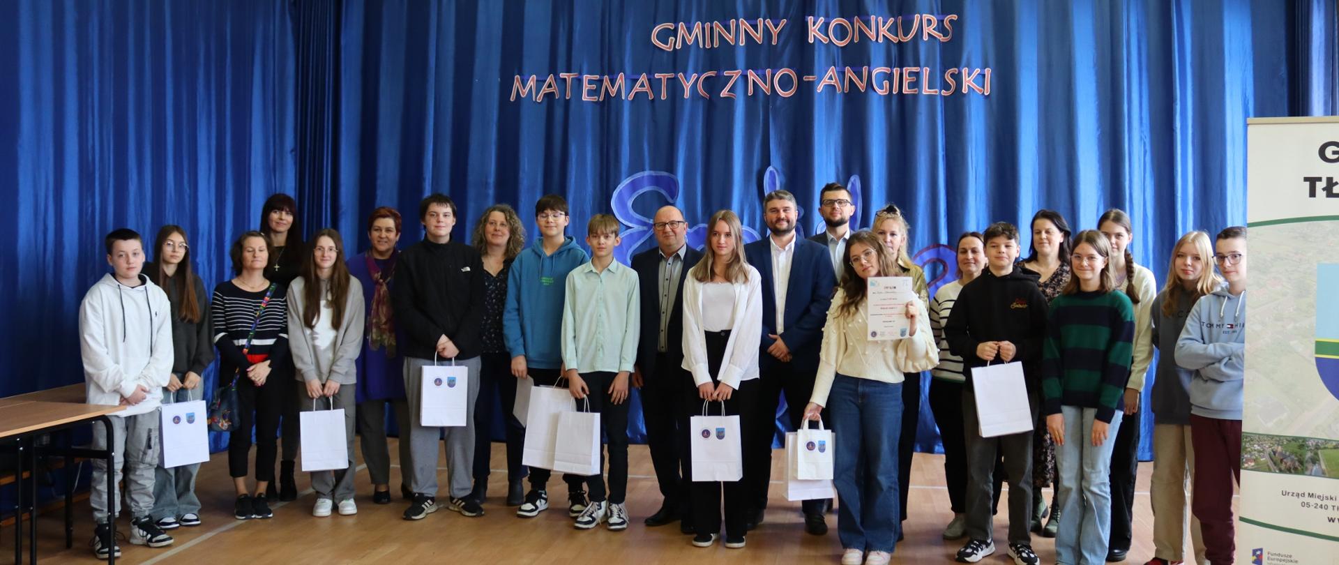 Grupa ludzi stojących obok siebie. Młodzież stojąca z przodu trzyma w rękach białe torebki. Za nimi niebieski materiał na ścianie z napisem gminny konkurs matematyczno angielski
