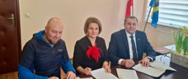 Na zdjęciu widoczne trzy osoby, siedzące za biurkiem z dokumentami. W tle ściana z obrazem i dwie flagi.