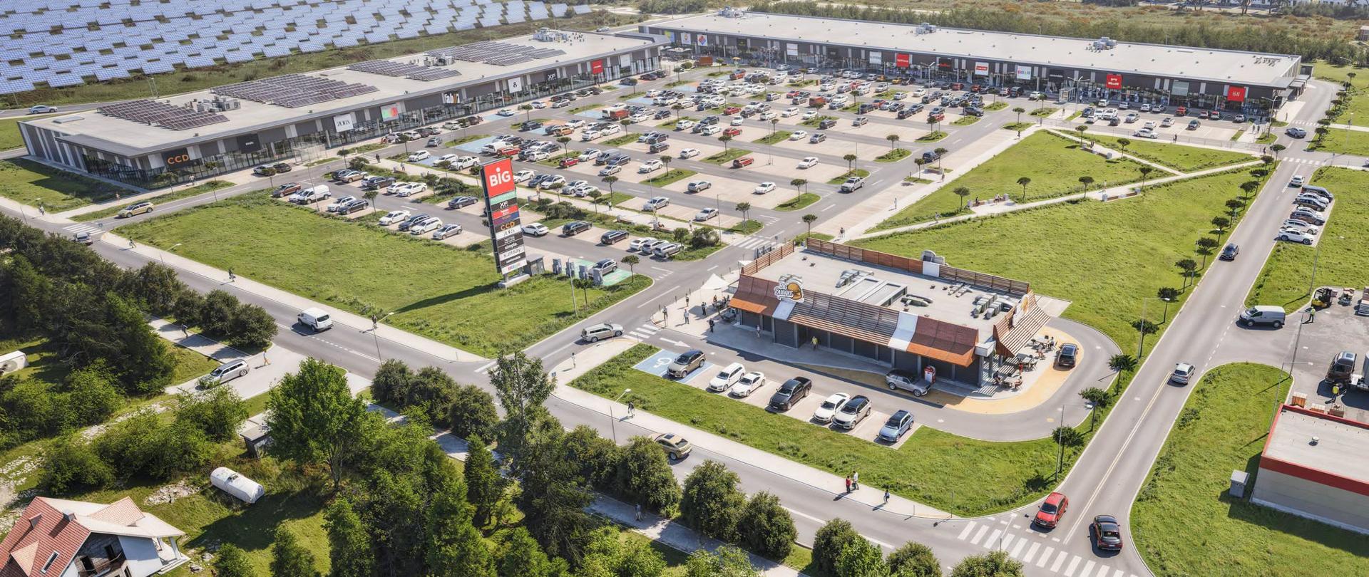 Wizualizacja kompleksu handlowo-usługowego z lotu ptaka. Widoczne są budynki sklepów, rozległy parking z samochodami, drogi dojazdowe, tereny zielone oraz instalacja fotowoltaiczna w sąsiedztwie obiektu.