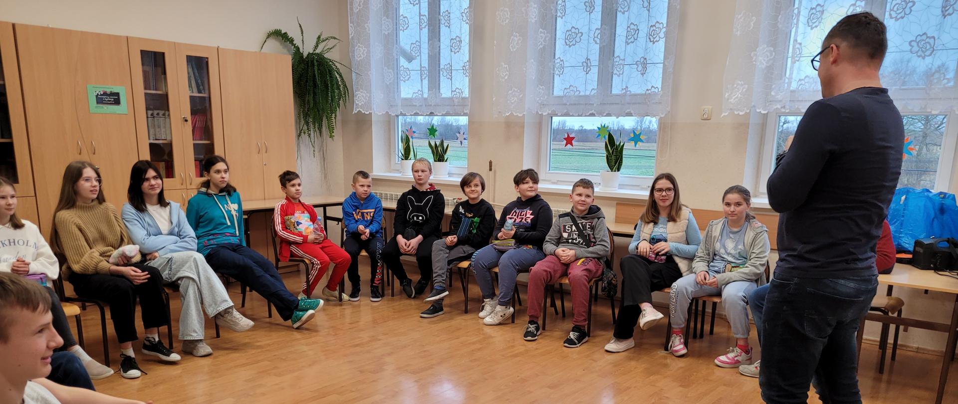 Uczniowie w sali lekcyjnej siedzą w kręgu, przed nimi stoi prowadzący warsztaty.