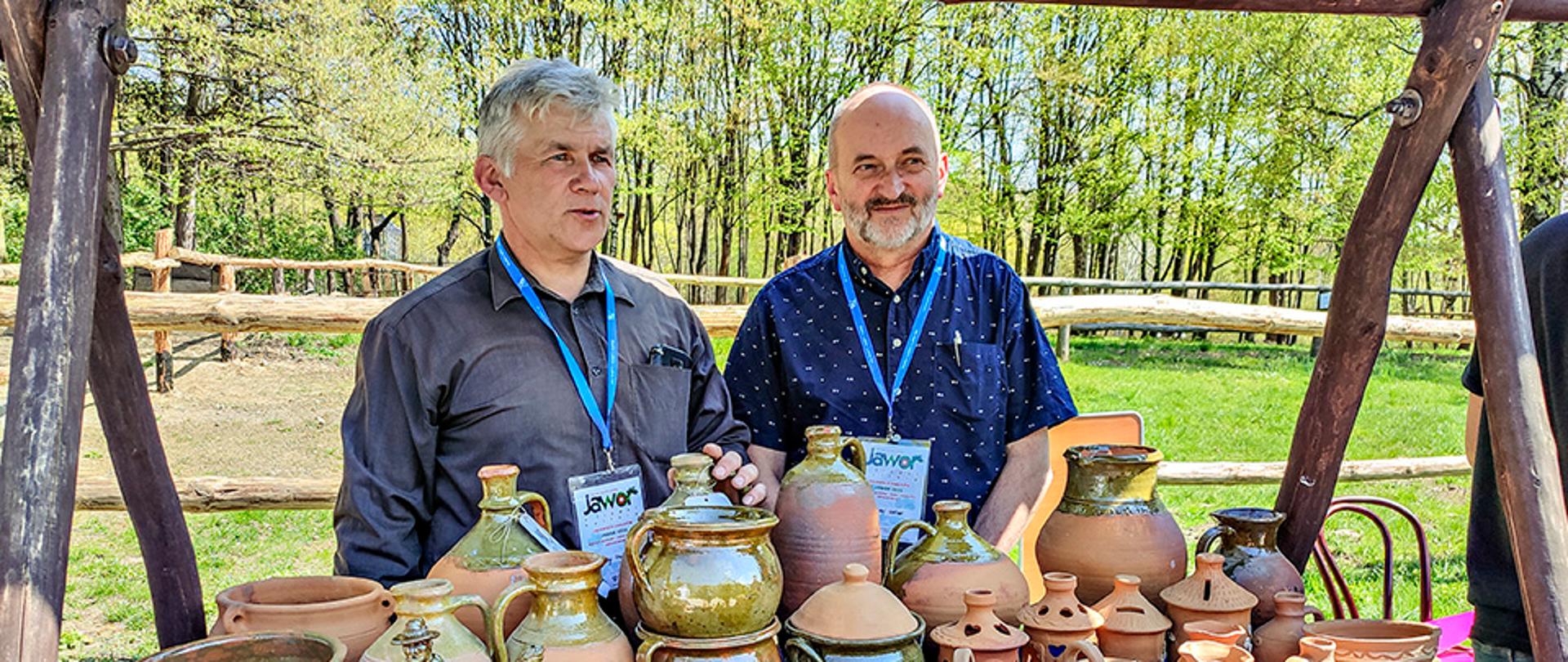 Na perwszym planie zdjęcia znajduje się stół przykryty niebieską tkaniną, na której ustawione zostały wyroby ceramiczne. Za stołem stoją dwie osoby. Od lewej Cezary Gajewski oraz Andrzej Gotner. W tle za nimi znajduje się zagroda z drewna otoczona drzewami.