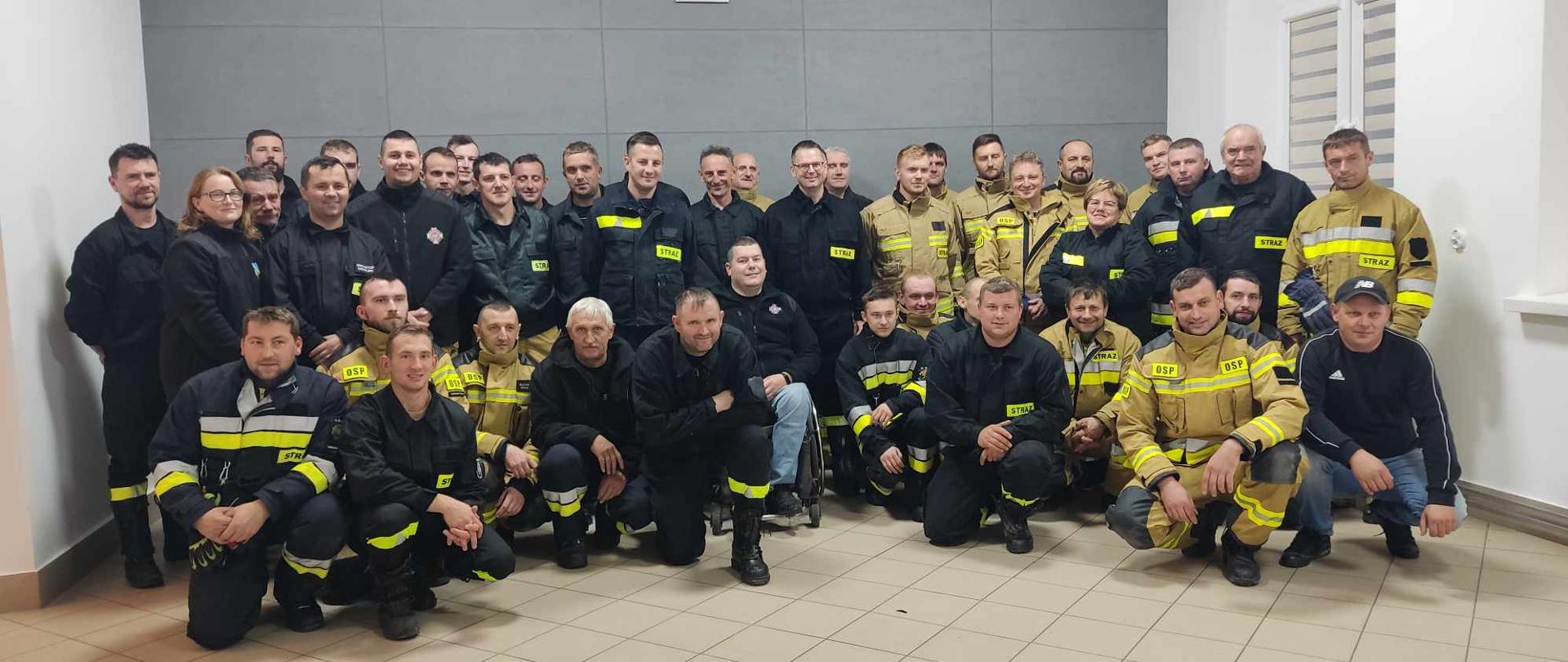 Zdjęcie przedstawia mężczyzn i kobiety w wieku od 20 do 65 lat, ubranych w stroje strażackie bojowe, pozujących w pomieszczeniu do zdjęcia.