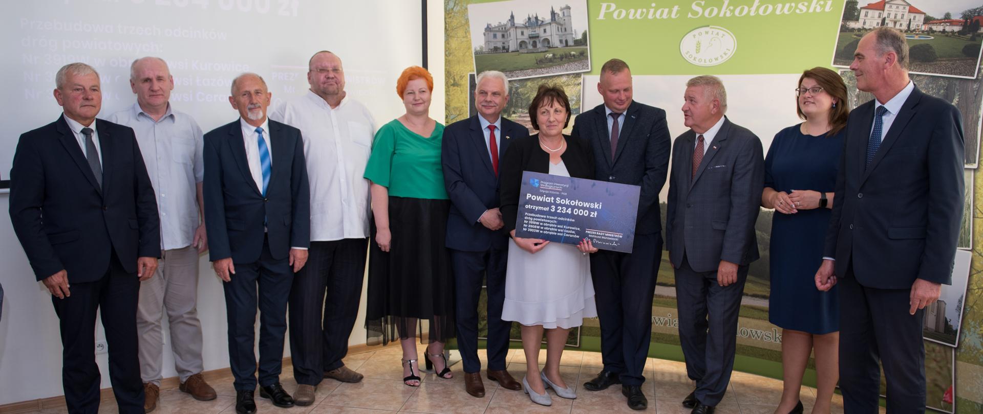 Zdjęcie grupowe reprezentacji Powiatu Sokołowskiego i moment wręczenia przez przedstawicieli rządu symbolicznego czeku na realizację inwestycji. Czek trzyma w dłoniach stojąca pośrodku starosta Elżbieta Sadowska.