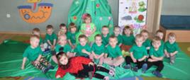 Dzieci ubrane w zielone koszulki siedzą na dywanie i zielonym materiale, przed nimi leży dziewczynka ubrana w strój biedronki