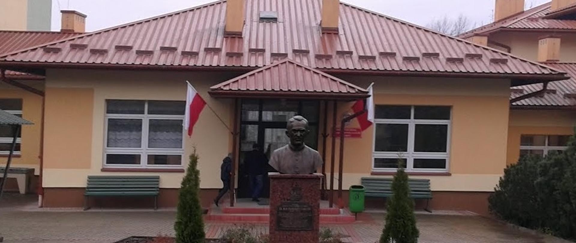 widok na pomnik patrona w tle wejście główne do szkoły wywieszone flagi państwowe obok pomnika i przed nim widoczna kostka brukowa i nasadzenia