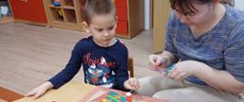 Chłopczyk i jego mama siedzą przy stole, wspólnie tworząc świąteczne ozdoby - papierową torbę z bożonarodzeniowym akcentem