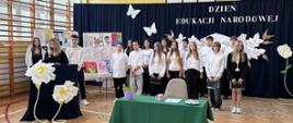 Grupa uczniów w galowych strojach stoi na tle granatowej kurtyny z napisem „DZIEŃ EDUKACJI NARODOWEJ” i papierowymi motylami. Po lewej stronie kobieta w czarnym stroju przemawia do mikrofonu przy mównicy ozdobionej dużymi, białymi papierowymi kwiatami na stojakach. W tle widoczne są tablice z pracami plastycznymi dzieci oraz stół z prezentami.