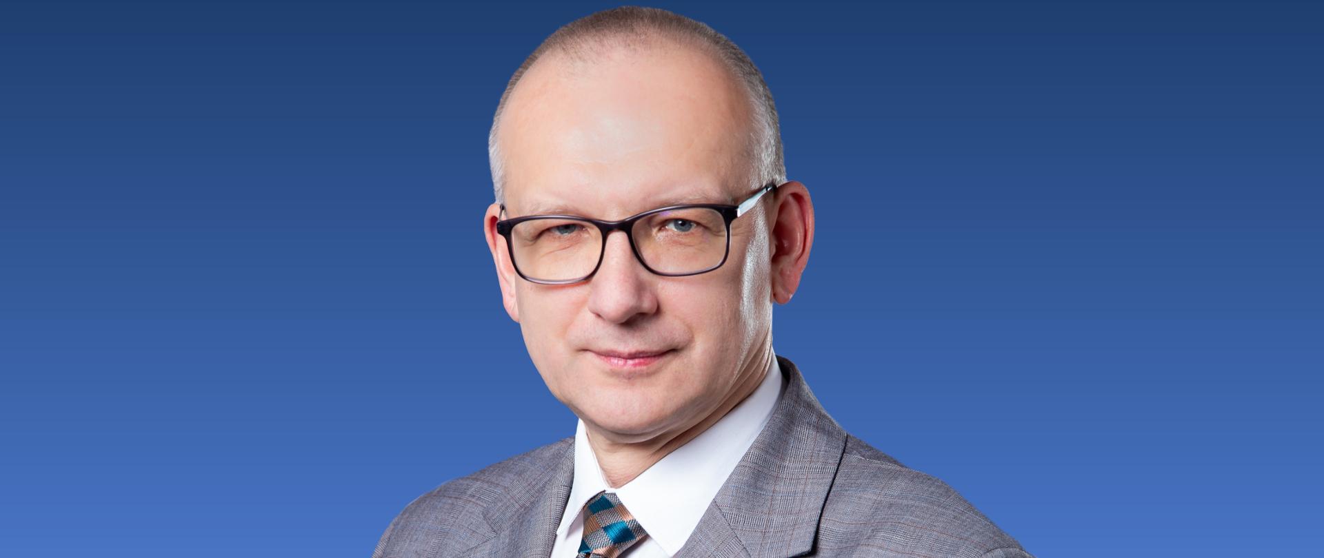 Zdjęcie przedstawia mężczyznę w okularach i w krawacie na niebieskim tle.