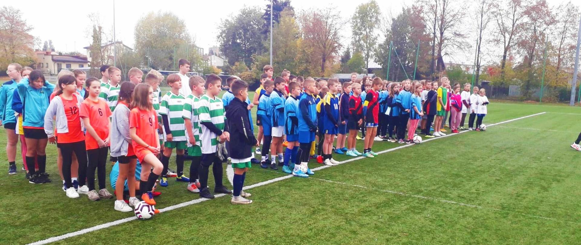 Zdjęcie przedstawia chłopców i dziewczęta podzielonych na drużyny, stojących na linii jednego z boisk Ośrodka Sportu i Rekreacji w Przasnyszu. Młodzież ze szkół podstawowych powiatu przasnyskiego bierze udział w powiatowych finałach w piłce nożnej w ramach Powiatowych Igrzysk Młodzieży Szkolnej.