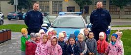 Policjanci stoją przy radiowozie a przed nimi grupa małych dzieci