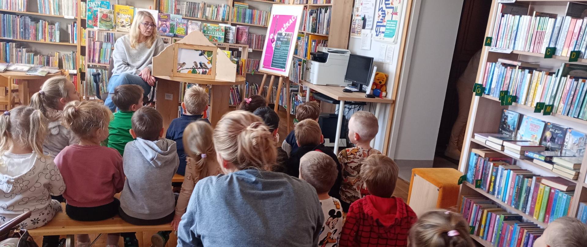 Przedszkolaki na załączonym obrazku słuchają bajki, którą czyta pani bibliotekarka