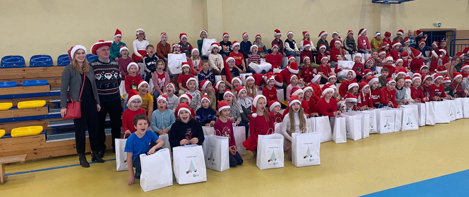 dzieci w czerwono-białych strojach świątecznych oraz 2 osoby dorosłe pozujące do zdjęcia na hali sportowej