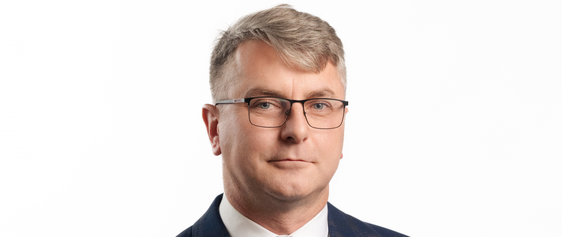 Siwiejący mężczyzna w okularach, jasne śmiejące się oczy. Ubrany w granatowy garnitur w szarą kratę białą koszulę i granatowy krawat w białe kropki. 