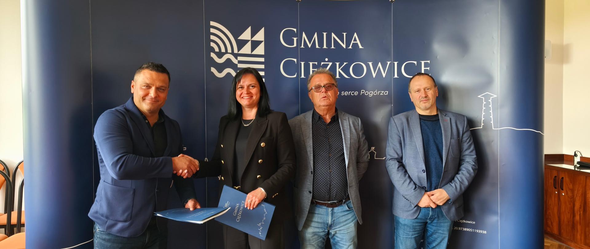 Cztery osoby stoją na tle niebieskiej ścianki z napisem 'Gmina Ciężkowice – Tu bije serce Pogórza'. Dwie osoby po lewej stronie (mężczyzna i kobieta) podają sobie ręce, trzymając niebieskie teczki z logo gminy. Po prawej stronie stoją dwaj inni mężczyźni w garniturach, również patrzący w stronę aparatu.