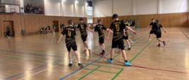 Chłopcy biorący udział w turnieju w czarnych koszulkach ze złotymi napisami wykonują rozgrzewkę na sali gimnastycznej