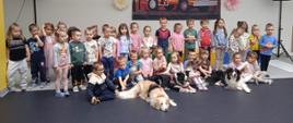 Grupa dzieci w jasnej sali pozuje do zdjęcia przed nimi na podłodze leżą psy
