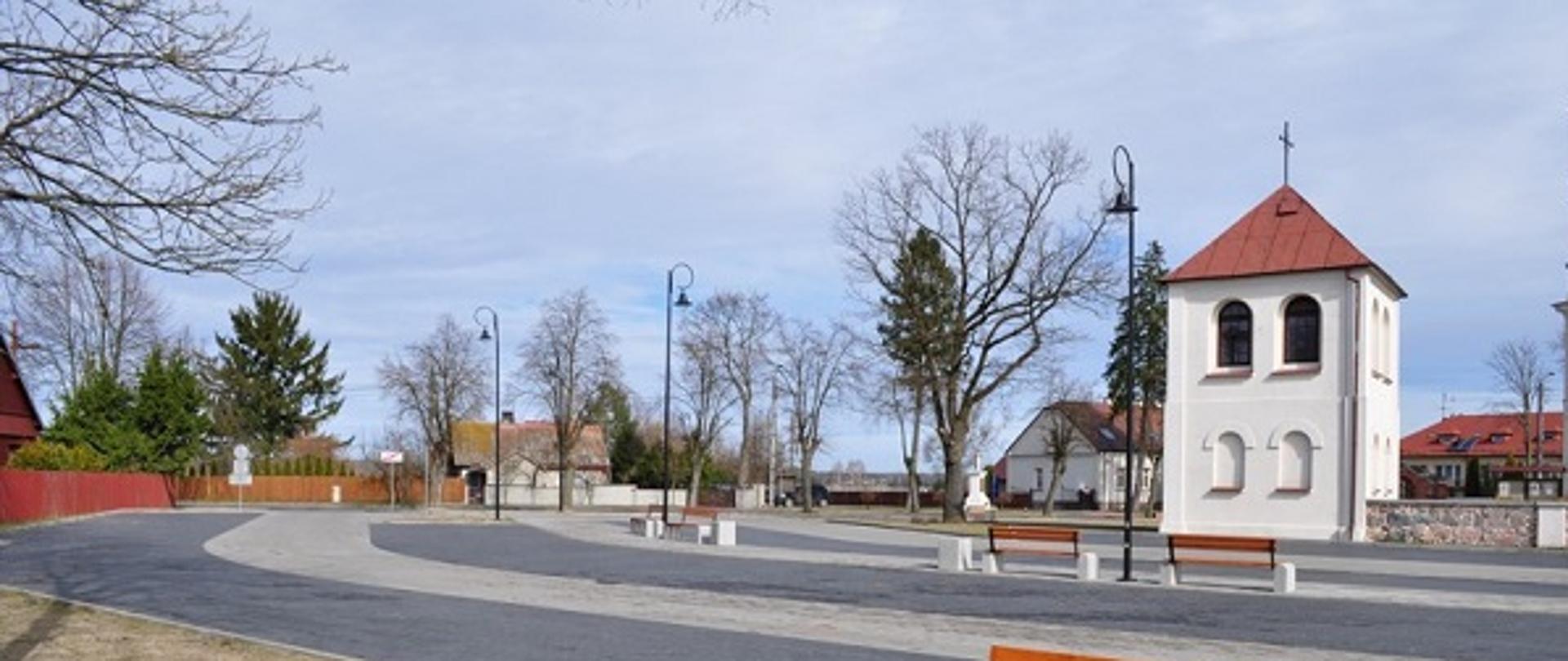Na zdjęciu widoczny jest wybudowany plac do rekreacji w pobliżu kościoła w miejscowości Poręba. Teren został wyłożony kostką brukową. Zamontowane są ławki, kosze na śmieci oraz latarnie. W tle znajduje się dzwonnica przy kościele w Porębie oraz inne zabudowania.