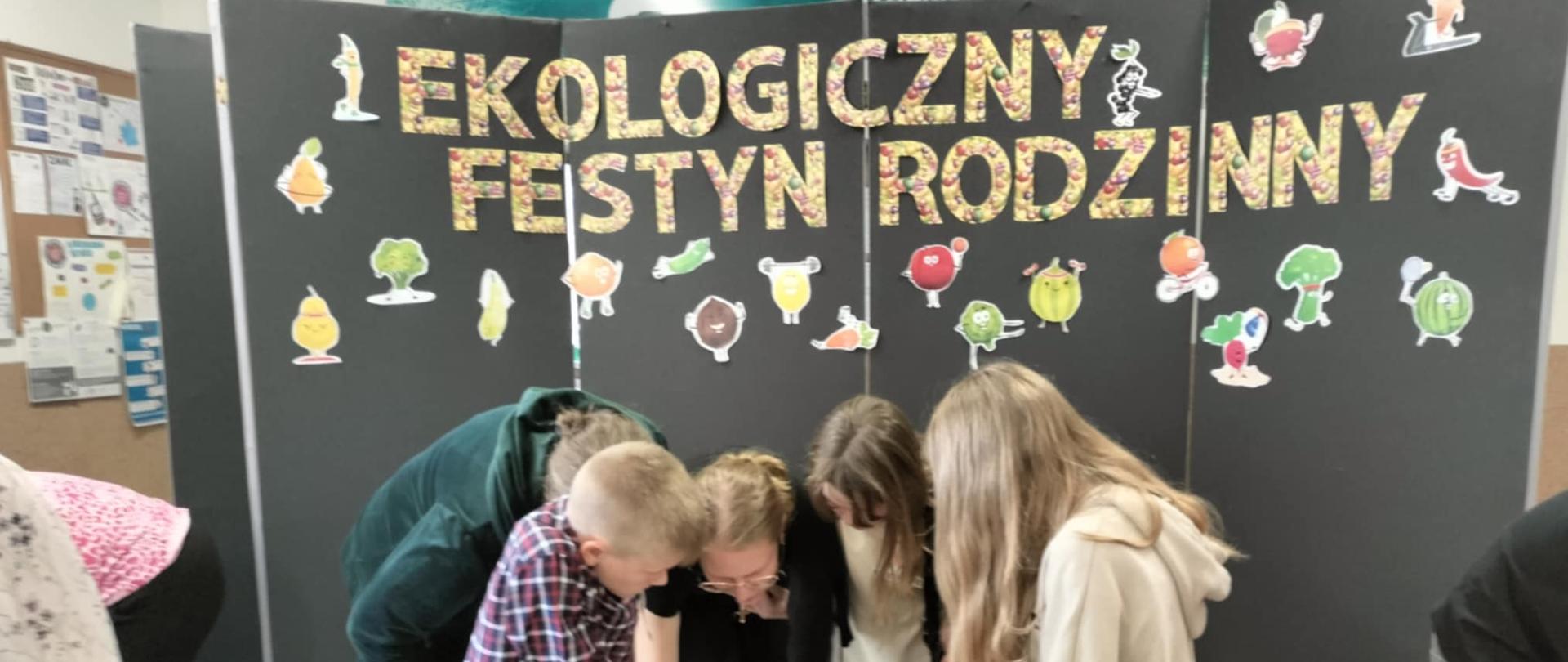 Na zdjęciu znajduje się czworo uczniów i dwie kobiety. Wszyscy pochylają się nad stolikiem, na którym leży kartka. Jedna z dziewczyn pisze po kartce.