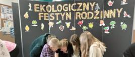 Na zdjęciu znajduje się czworo uczniów i dwie kobiety. Wszyscy pochylają się nad stolikiem, na którym leży kartka. Jedna z dziewczyn pisze po kartce.