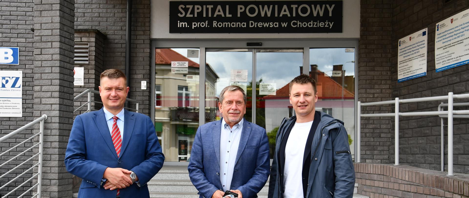 3 osoby stojące przed wejściem do budynku, nad wejściem napis Szpital Powiatowy im. prof. Romana Drewsa w Chodzieży