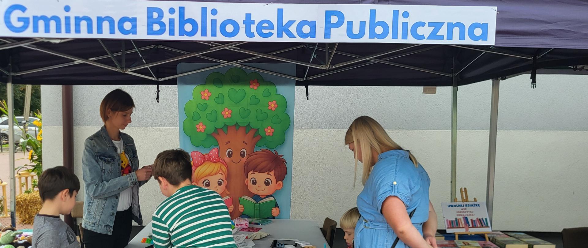 Pod namiotem przy stole stoją dzieci oraz dwie kobiety. Jedna z kobiet przygląda się siedzącemu przy stole chłopcu. Chłopiec w ręku trzyma flamaster i ozdabia białą bawełnianą czapkę.