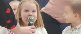 Przedszkolaki stoją w sali na tle ścianki "Pasowanie na przedszkolaka". Przy mikrofonie stoi jeden z przyszłych przedszkolaków.