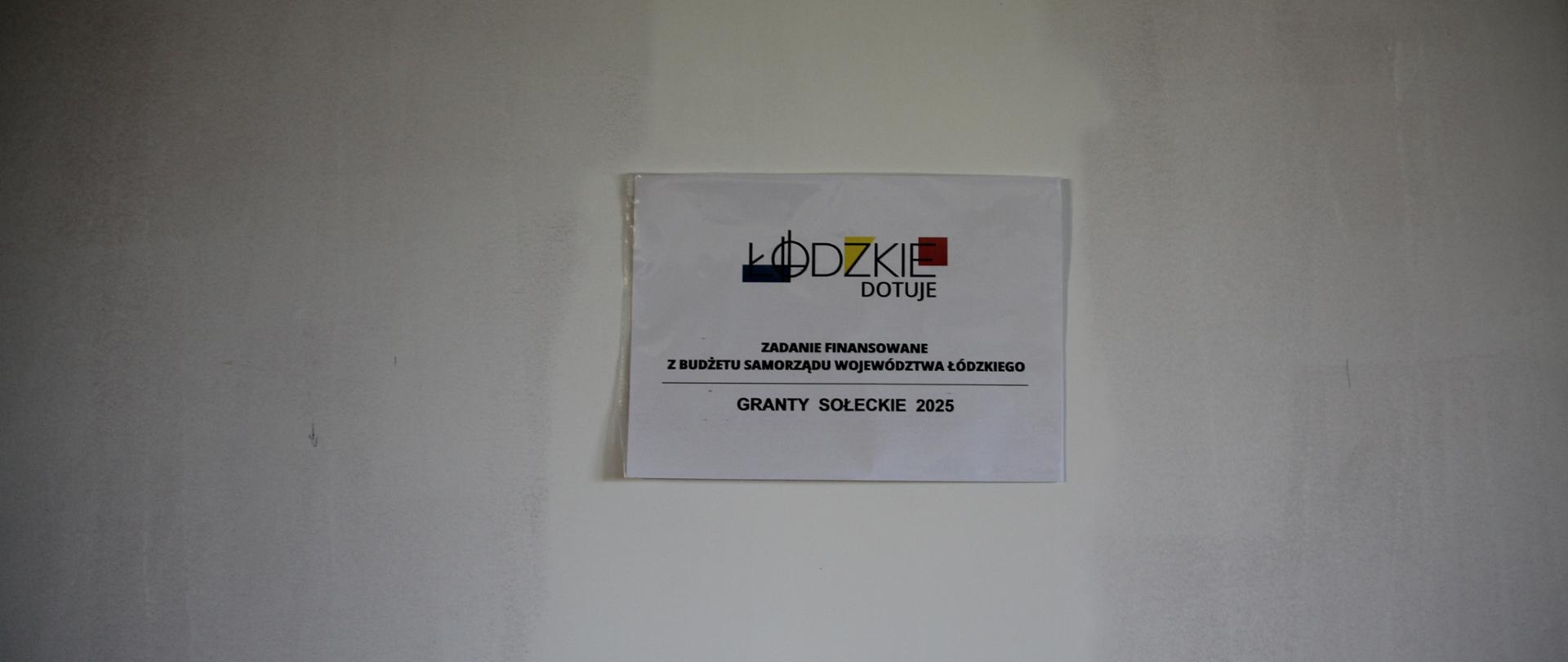 Zbliżenie na białą kartkę z logo "ŁÓDZKIE DOTUJE" i tekstem w języku polskim, umieszczoną centralnie na jasnej, nieukończonej ścianie. Na kartce widnieje informacja, że zadanie jest finansowane z budżetu Samorządu Województwa Łódzkiego w ramach programu "GRANTY SOŁECKIE 2025". Tło to jednolita, surowa ściana.