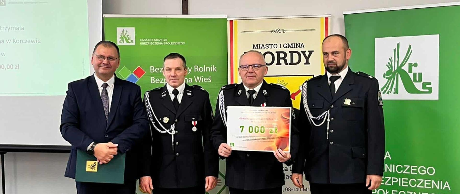 cztery osoby- w tym trzy w galowych mundurach strażackich. Jedna z nich trzyma tablicę- czek na 7000zł. Za nimi tablice: KRUS, Gminy Mordy.