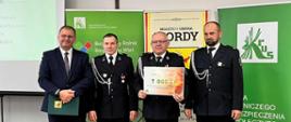 cztery osoby- w tym trzy w galowych mundurach strażackich. Jedna z nich trzyma tablicę- czek na 7000zł. Za nimi tablice: KRUS, Gminy Mordy.