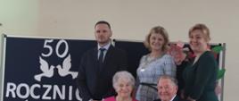 Państwo Irena i Henryk na tle granatowej tablicy z napisem wraz z Wójtem, Z-cą Wójta i Z-cą Kierownika_USC